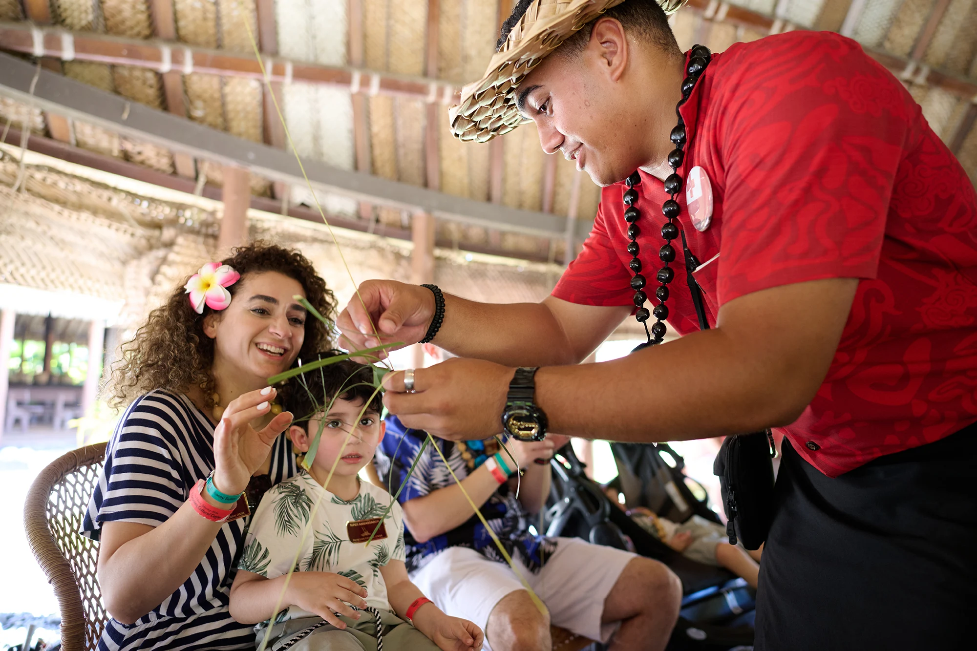 guide in a red shirt and woven hat weaves a palm leaf headband for a smiling young boy, as his mother, wearing a plumeria flower in her curly hair, watches and helps