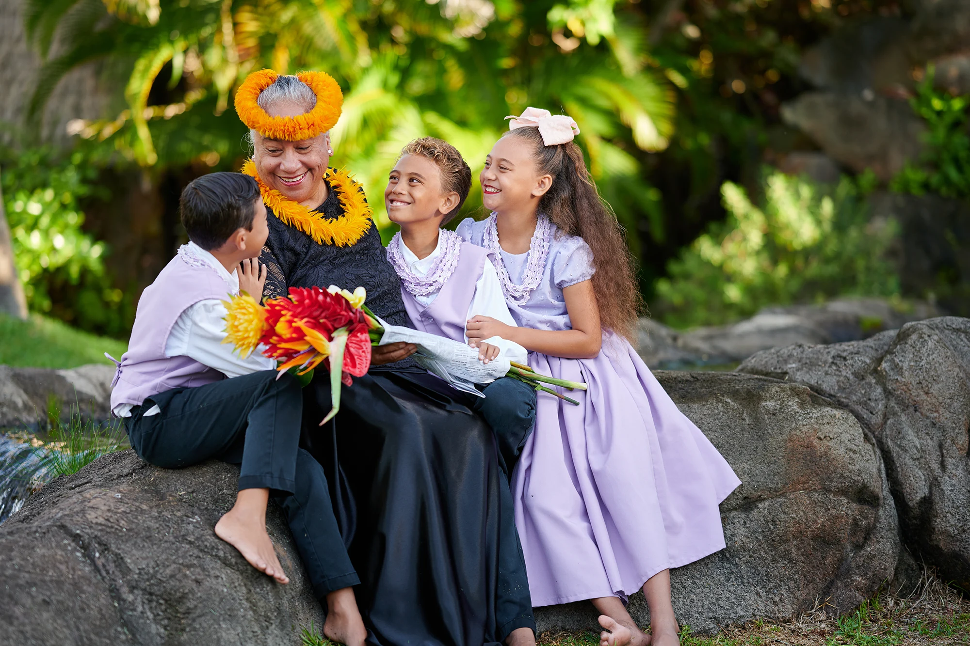 A smiling woman wearing a yellow flower crown and lei sits on a rock with three children in lavender and white traditional Hawaiian outfits, holding tropical flowers, in a lush garden setting in Hawai'i.
