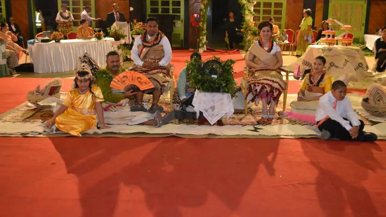 Adults and children sit on woven mats at a formal wedding ceremony, wearing traditional Tongan attire with flower garlands and patterned garments.