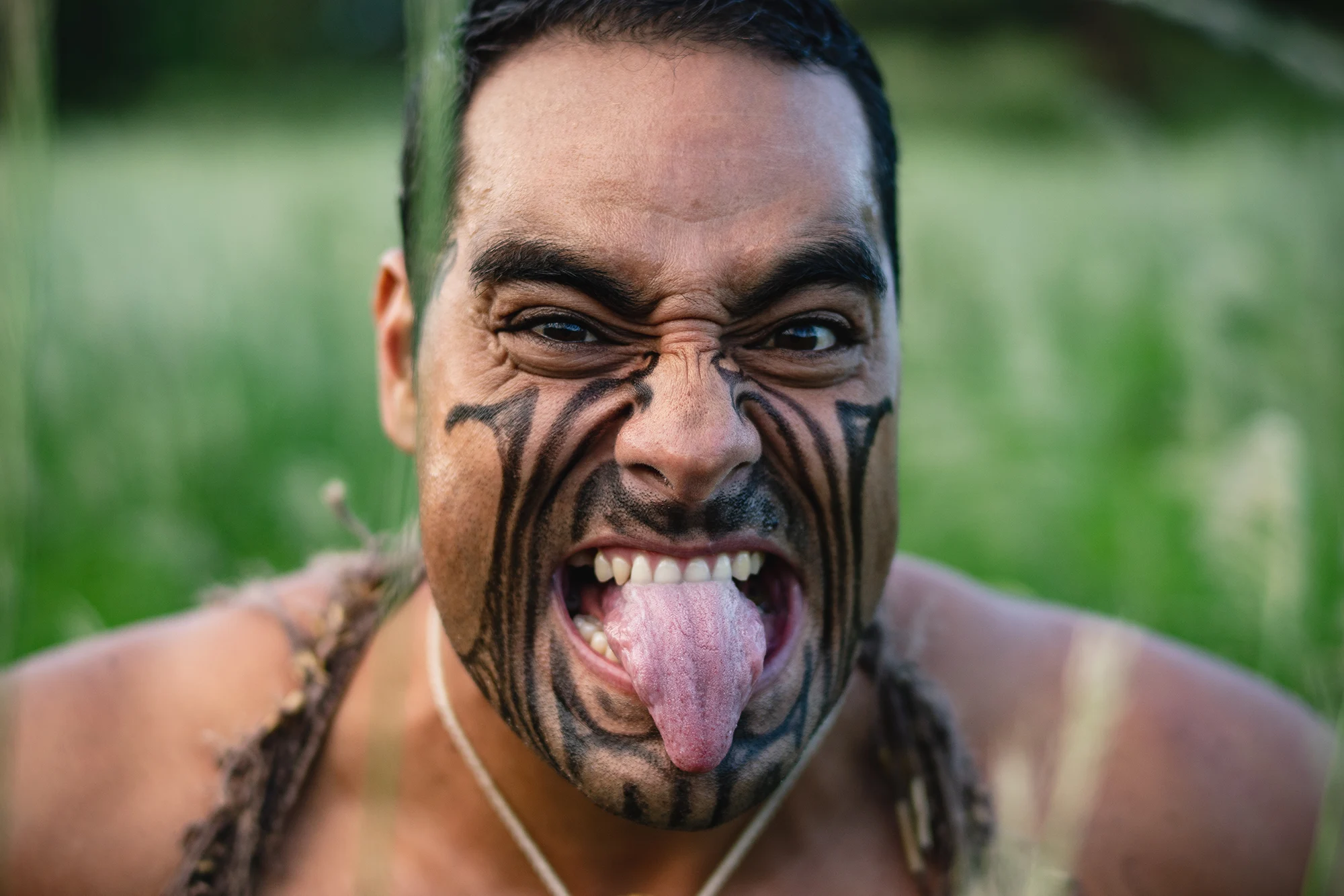 close-up of a Māori performer with facial tā moko showing wide eyes and extended tongue during a haka in a grassy field.
