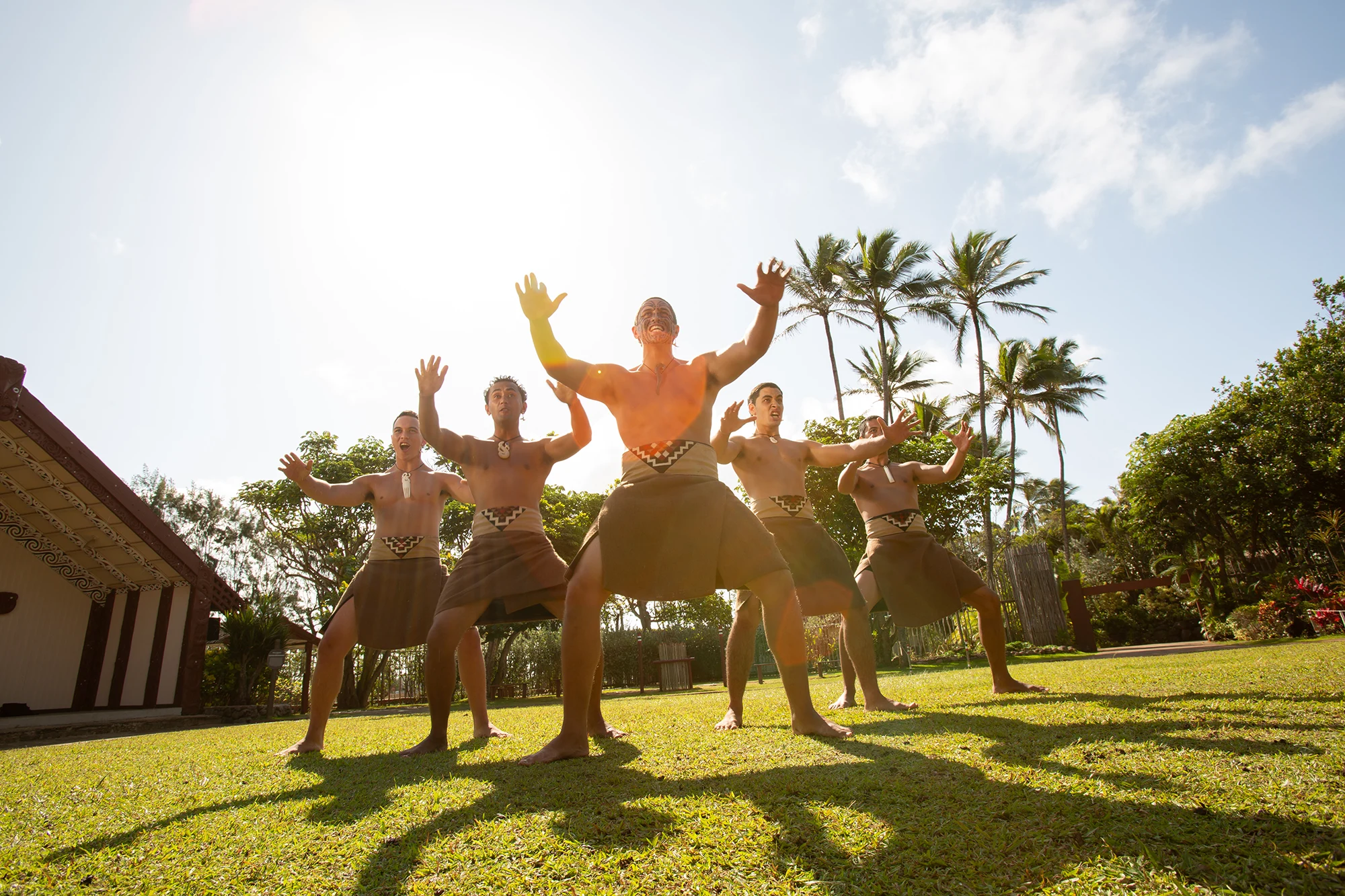 Five Māori performers in traditional attire raise their arms and perform the haka on a sunlit lawn, with a carved marae and palm trees in the background at the Polynesian Cultural Center.