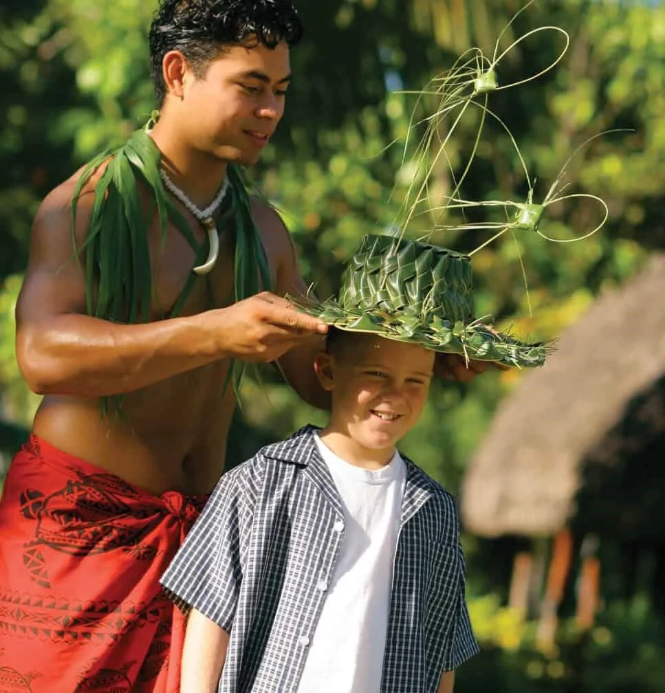 Samoan host places coconut leaf hat on young guest.
