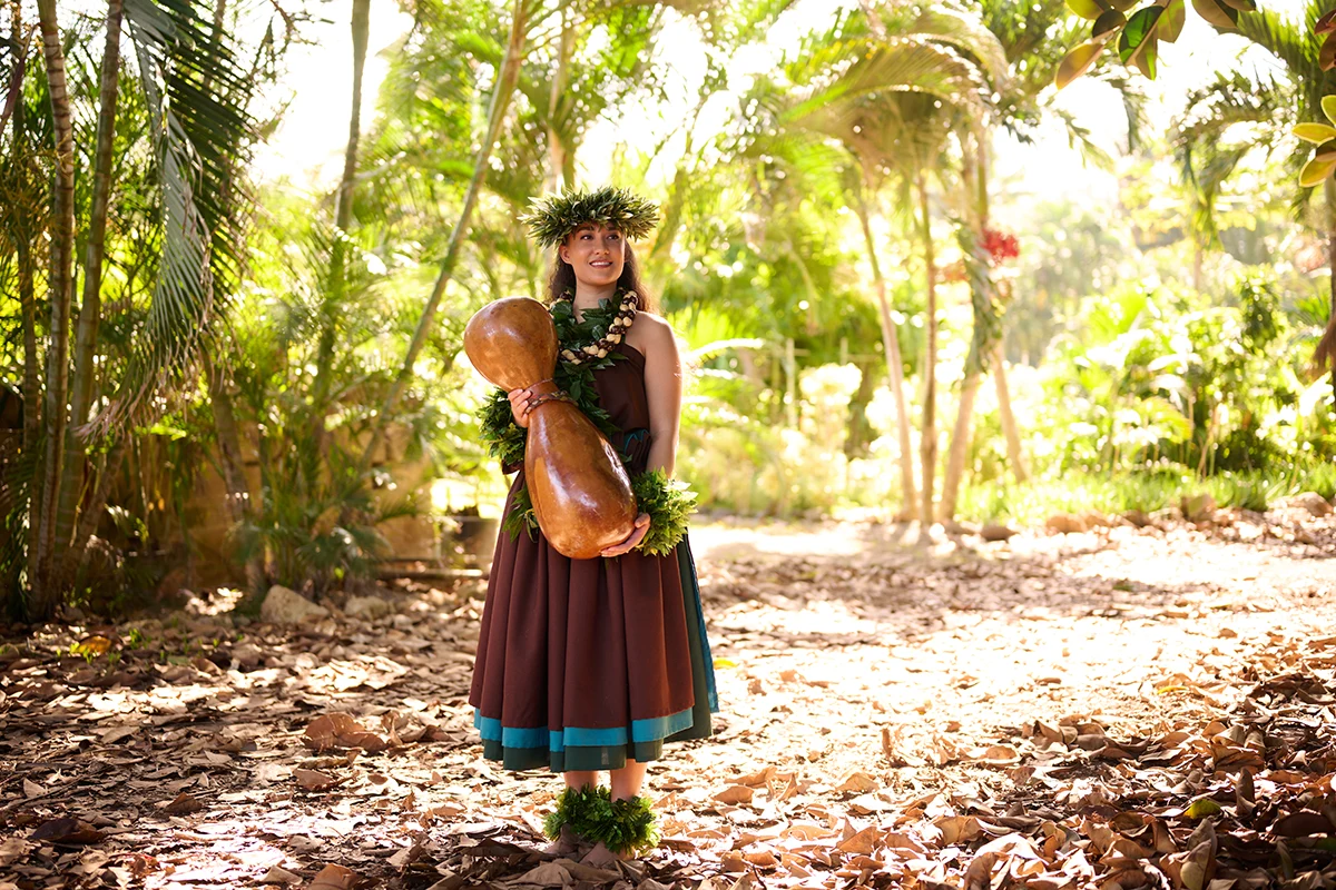 A Hawaiian island villager stands in a tropical forest, holding a traditional ipu (gourd instrument) amid lush green foliage.