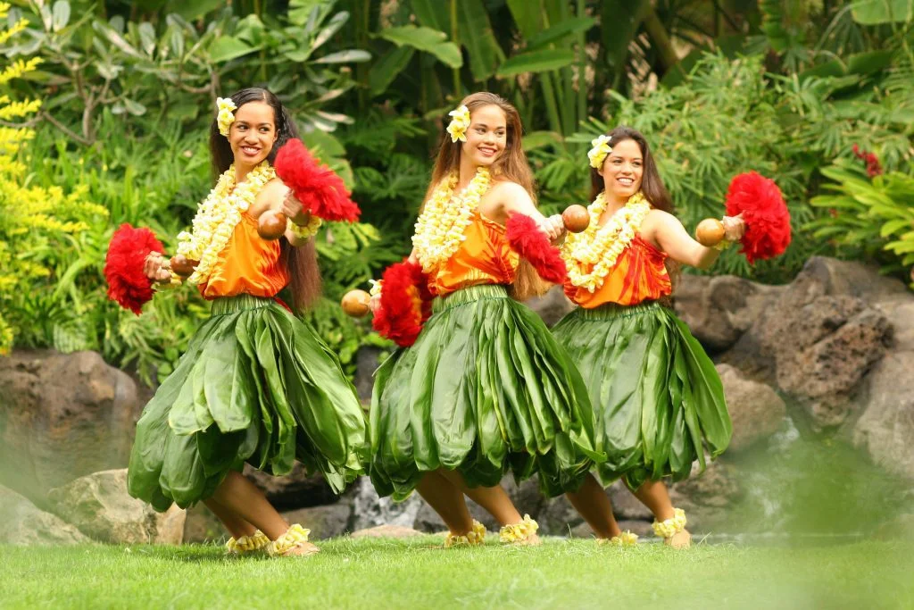 Three Polynesian women dressed in tī leaves, leis and holding uli uli implements