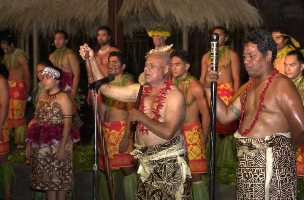 A Samoan elder dressed in traditional attire speaks into a microphone. Several men and women, all dressed in traditional Samoan attire, stand behind him.